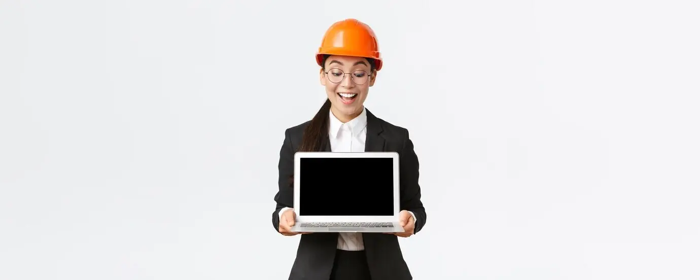 An impressed, happy Asian female chief engineer, wearing a safety helmet and business suit, stands holding a laptop, presenting the company’s great results to investors and looking pleased at the computer screen.