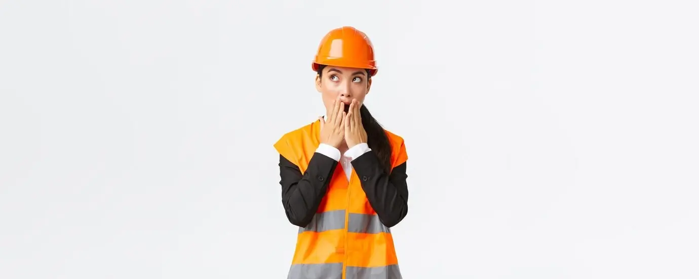 Shocked and concerned Asian female engineer-architect in a safety helmet and reflective jacket, looking toward the upper left corner and gasping with a worried, amazed stare, standing against a white background.